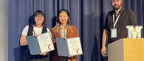 Die Preisträgerinnen Simona Bianco und Jiarong WU stehen auf der Bühne mit ihren Urkunden neben dem Schatzmeister der RSC MASC, Dr. Andrew Surman